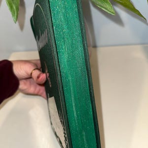 Puede incluir: Un libro de tapa dura con una cubierta verde oscuro y bordes verdes brillantes. La cubierta del libro presenta texto blanco y un dise&ntilde;o circular. El libro est&aacute; sostenido por una mano, con hojas verdes en el fondo.