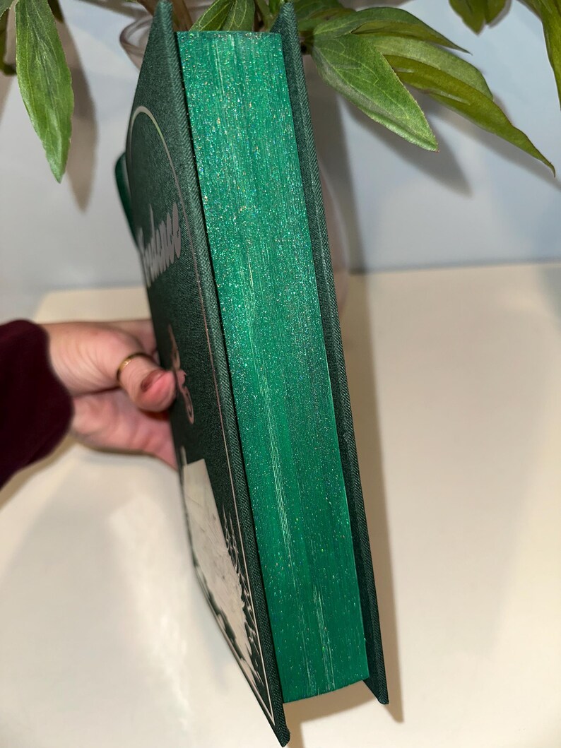 Puede incluir: Un libro de tapa dura con una cubierta verde oscuro y bordes verdes brillantes. La cubierta del libro presenta texto blanco y un dise&ntilde;o circular. El libro est&aacute; sostenido por una mano, con hojas verdes en el fondo.