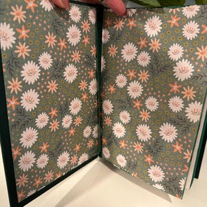 Puede incluir: Diario abierto con una cubierta verde oscuro y p&aacute;ginas con estampado floral. Las p&aacute;ginas interiores presentan un patr&oacute;n repetido de flores blancas y melocot&oacute;n sobre un fondo verde apagado. El diario est&aacute; abierto, mostrando el dise&ntilde;o detallado.