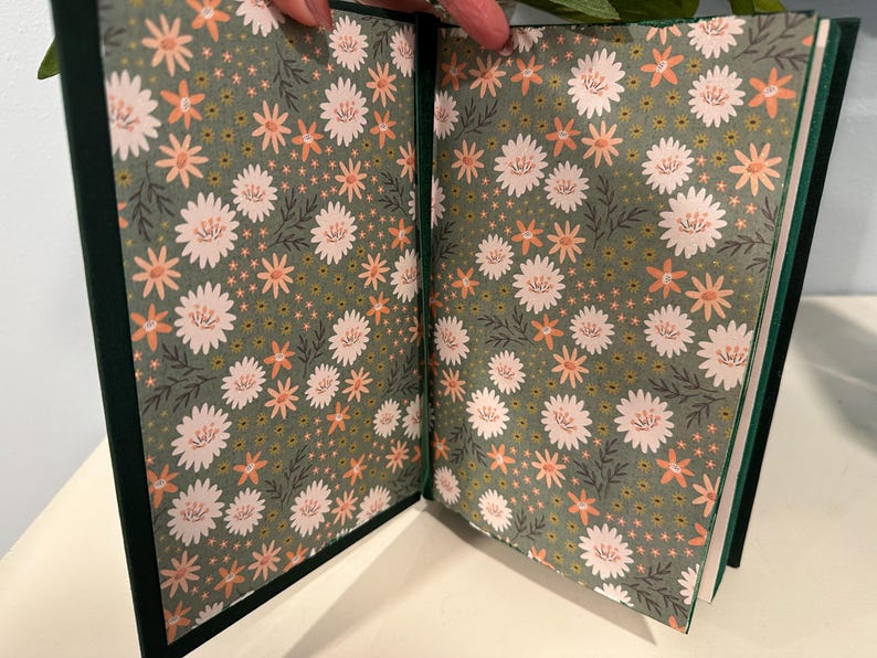 Puede incluir: Diario abierto con una cubierta verde oscuro y p&aacute;ginas con estampado floral. Las p&aacute;ginas interiores presentan un patr&oacute;n repetido de flores blancas y melocot&oacute;n sobre un fondo verde apagado. El diario est&aacute; abierto, mostrando el dise&ntilde;o detallado.