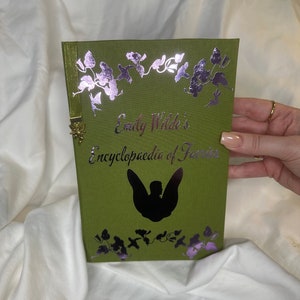 May include: A green book with a silver foil design of flowers and a fairy silhouette. The title is "Emily Wilde's Encyclopaedia of Faeries" in pink and black lettering.