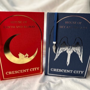 Puede incluir: Dos libros de tapa dura, uno rojo y otro azul marino, con letras y diseños dorados y plateados. El libro rojo dice "House of Earth and Blood" y "Crescent City". El libro azul dice "House of Sky and Breath" y "Crescent City."