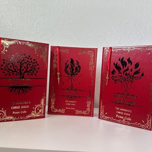 Puede incluir: Tres libros de tapa dura rojos con detalles dorados. Cada libro presenta un gráfico negro único y letras doradas. Se adjuntan marcadores de cinta roja con dijes dorados. Los títulos incluyen "The Kindred's Curse Saga" y "Heat of the Everflame".