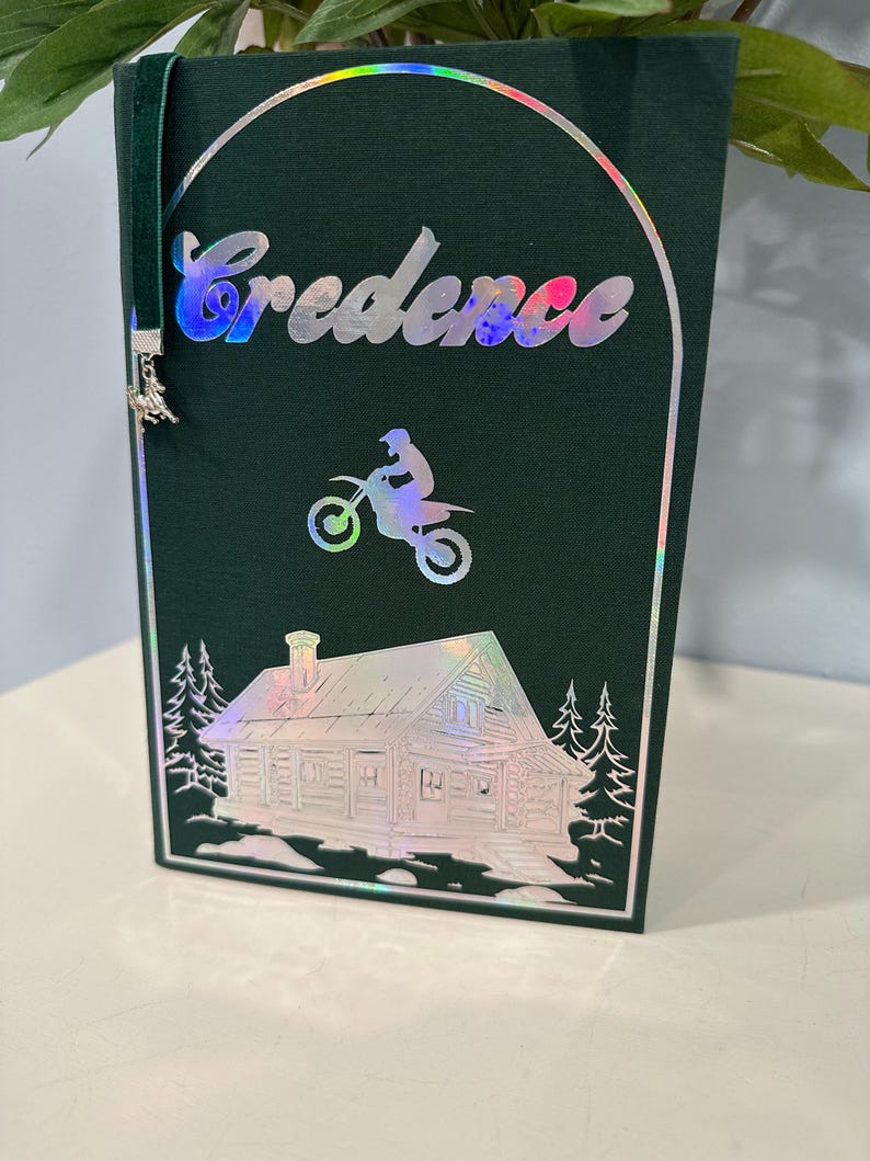 Puede incluir: Cubierta de libro verde oscuro con la palabra "Credence" en letras hologr&aacute;ficas. Tambi&eacute;n se presenta una imagen hologr&aacute;fica de una persona en una motocicleta y una caba&ntilde;a. Se adjunta un marcap&aacute;ginas de terciopelo verde con un dije.
