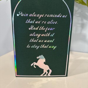 Puede incluir: Una tarjeta de felicitaci&oacute;n verde oscuro con un borde hologr&aacute;fico y un borde blanco. La tarjeta presenta el texto "Pain always reminds us that we're alive. And the fear along with it that we want to stay that way." Una silueta de caballo rosa y azul est&aacute; en la parte inferior.