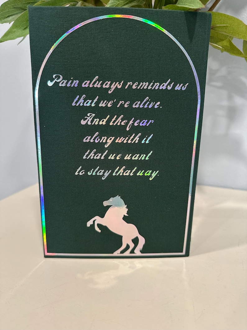 Puede incluir: Una tarjeta de felicitaci&oacute;n verde oscuro con un borde hologr&aacute;fico y un borde blanco. La tarjeta presenta el texto "Pain always reminds us that we're alive. And the fear along with it that we want to stay that way." Una silueta de caballo rosa y azul est&aacute; en la parte inferior.