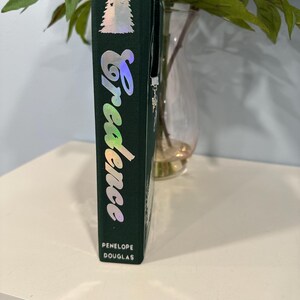 Puede incluir: Un libro de tapa dura verde oscuro con el t&iacute;tulo "Credence" en fuente cursiva hologr&aacute;fica. El nombre del autor, Penelope Douglas, est&aacute; impreso en la parte inferior. Un dise&ntilde;o de &aacute;rbol plateado est&aacute; en la parte superior.