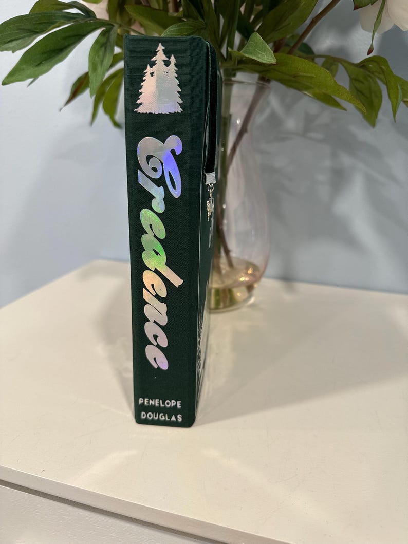 Puede incluir: Un libro de tapa dura verde oscuro con el t&iacute;tulo "Credence" en fuente cursiva hologr&aacute;fica. El nombre del autor, Penelope Douglas, est&aacute; impreso en la parte inferior. Un dise&ntilde;o de &aacute;rbol plateado est&aacute; en la parte superior.