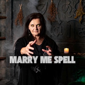 May include: A woman with glowing yellow eyes and dark hair, wearing a black outfit, is the focus of this image. The text "MARRY ME SPELL" is displayed in white across the bottom. A candle and decorative elements are in the background.