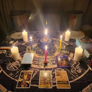 Op de afbeelding: Een donkerblauw tafelkleed met een cirkelvormig ontwerp met astrologische symbolen en tarotkaarten. Zeven brandende kaarsen zijn rond de tafel geplaatst, met een roze, gele en blauwe kaars in het midden.