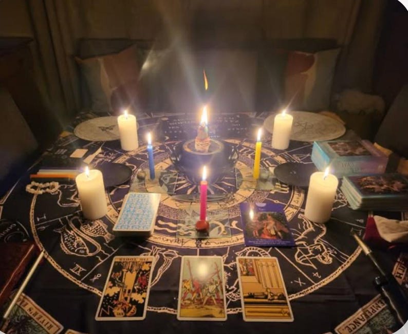 Op de afbeelding: Een donkerblauw tafelkleed met een cirkelvormig ontwerp met astrologische symbolen en tarotkaarten. Zeven brandende kaarsen zijn rond de tafel geplaatst, met een roze, gele en blauwe kaars in het midden.