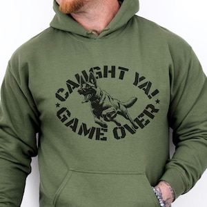 May include: Olive green hooded sweatshirt with a front pocket. The sweatshirt features a black graphic of a running dog inside a circle with the words "CAUGHT YA! GAME OVER" in a bold font.