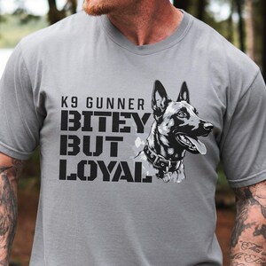 May include: Gray t-shirt featuring a black and white illustration of a dog with the text "K9 Gunner, Bitey But Loyal". The dog is wearing a collar. The shirt is a crew neck.