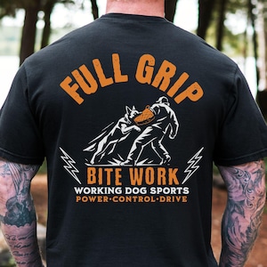 May include: A black short-sleeve t-shirt featuring a graphic on the back. The design shows a dog biting a training sleeve held by a person, with the text FULL GRIP BITE WORK, WORKING DOG SPORTS, and POWER CONTROL DRIVE in orange and white.