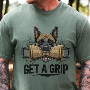 May include: Olive green t-shirt featuring a graphic of a German Shepherd dog biting a wooden training dumbbell. The text "GET A GRIP" is printed in bold, black letters below the dog.