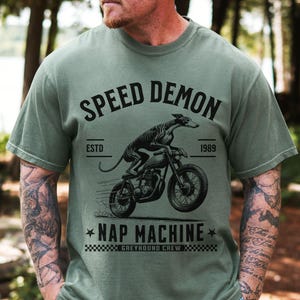 May include: A sage green short-sleeved t-shirt featuring a black graphic of a greyhound riding a motorcycle. The text reads SPEED DEMON, ESTD 1989, NAP MACHINE, and GREYHOUND CREW, accompanied by a checkered pattern and two small star icons.