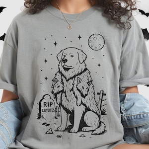May include: A gray t-shirt featuring a black graphic of a dog with three wolf heads, a tombstone that reads "RIP Coyotes", a moon, and stars. The shirt is a relaxed fit.