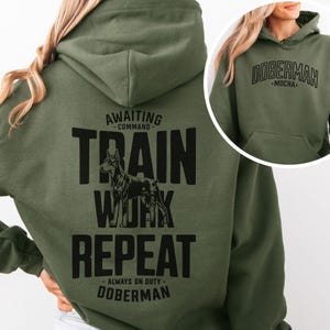 May include: Olive green hoodie with a front pocket and drawstring hood. The back of the hoodie features the text "Awaiting Command - Train Work Repeat - Always on Duty - Doberman" with a Doberman graphic. The front of the hoodie says "Doberman - Mocha."