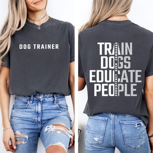 May include: A charcoal gray short-sleeve t-shirt featuring the white text DOG TRAINER on the front. The back displays the white text TRAIN DOGS EDUCATE PEOPLE with the vertical phrase CONSISTENCY OVER COMMANDS in a striped font style.