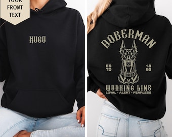 May include: A black hooded sweatshirt featuring the text HUGO on the front. The back displays a graphic of a Doberman dog with the text DOBERMAN, ESTD 1890, WORKING LINE, LOYAL, ALERT, FEARLESS in a light-colored font against the black fabric.