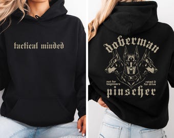 May include: A black hooded sweatshirt featuring the text tactical minded in a gothic font on the front. The back displays a graphic of three Doberman Pinscher heads with the text doberman pinscher, not for beginners, and rated D for drive.