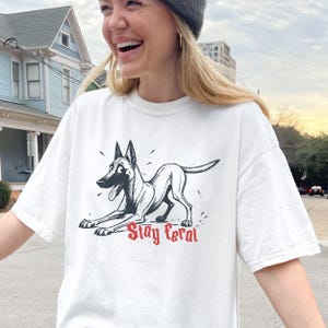 May include: A white short-sleeve t-shirt featuring a black ink-style illustration of a dog in a play bow position with its tongue out. Below the dog, the text Stay Feral is printed in a bold, red, gothic-inspired font on the front of the shirt.