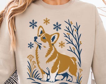 May include: A beige crewneck sweatshirt featuring a graphic illustration of a Corgi dog standing in snow. The design includes dark blue and gold snowflakes, bare branches, and floral elements surrounding the dog in a winter scene.