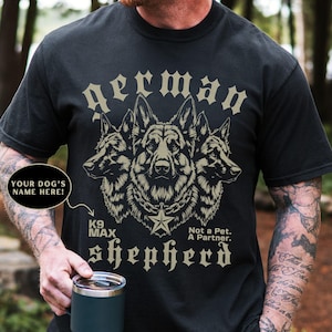 May include: Black t-shirt with a detailed illustration of three German Shepherds. The design includes the text "German Shepherd", "K9 MAX", and "Not a Pet. A Partner.". There is space to add your dog's name.
