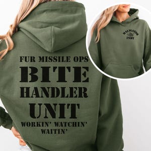 May include: Olive green hooded sweatshirt with the text "FUR MISSILE OPS BITE HANDLER UNIT WORKIN' WATCHIN' WAITIN'" on the back. The front of the sweatshirt has the text "MALIGATOR INDY". The sweatshirt has a front pocket and a hood.