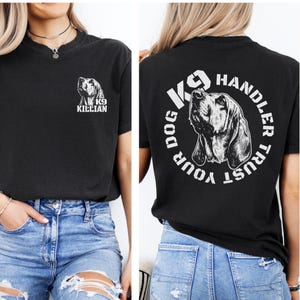 Puede incluir: Una camiseta negra con un gráfico blanco de una cabeza de perro en el pecho izquierdo con el texto K9 KILLIAN. La parte posterior de la camiseta muestra un gráfico blanco más grande de una cabeza de perro rodeado por el texto K9 HANDLER TRUST YOUR DOG.