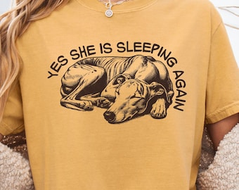 May include: A mustard yellow short-sleeve t-shirt featuring a dark brown graphic illustration of a sleeping greyhound dog. The text "YES SHE IS SLEEPING AGAIN" is printed in a dark brown arched font above the dog illustration on the chest.