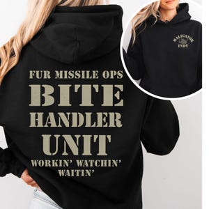 Puede incluir: Sudadera con capucha negra con el texto "FUR MISSILE OPS BITE HANDLER UNIT WORKIN' WATCHIN' WAITIN'" en la espalda. La parte delantera de la sudadera tiene el texto "MALIGATOR INDY". El texto es de color beige.
