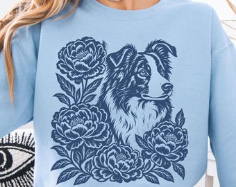 May include: A light blue crewneck sweatshirt featuring a dark navy blue graphic print on the front. The design displays a detailed illustration of a Border Collie dog surrounded by several large, blooming peony flowers and leaves.
