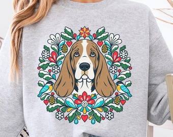 May include: A light gray crewneck sweatshirt featuring a central graphic of a brown and white Basset Hound dog surrounded by a colorful wreath of green leaves, red hearts, blue birds, pinecones, and white snowflakes on the front.
