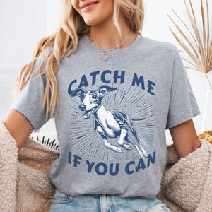May include: A heather grey short-sleeved t-shirt featuring a dark blue graphic of a running greyhound dog. The text CATCH ME IF YOU CAN is printed in dark blue capital letters above and below the dog illustration with radiating lines.
