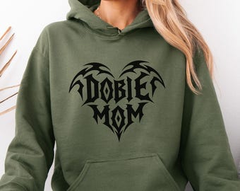 May include: An olive green hooded sweatshirt featuring a black graphic print on the chest. The design consists of the text DOBIE MOM arranged in a heart shape with sharp, pointed edges and tribal-style lettering. The hoodie has a front pocket.