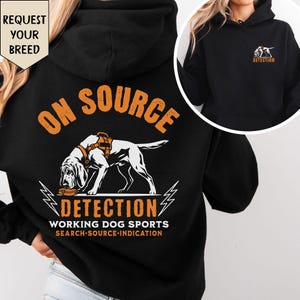 May include: A black hooded sweatshirt featuring a graphic of a dog in a harness sniffing a small case. The text reads ON SOURCE DETECTION WORKING DOG SPORTS SEARCH SOURCE INDICATION. A smaller version of the graphic is on the front chest.
