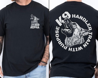 May include: A black t-shirt featuring a white graphic of a German Shepherd dog on the front left chest with the text K9 ALPHA. The back displays a larger version of the same dog graphic surrounded by the text K9 HANDLER TRAIN WITH PURPOSE.
