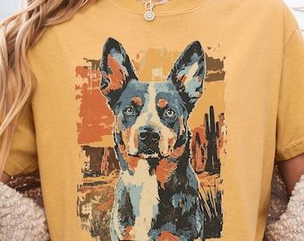 May include: Mustard yellow t-shirt featuring a graphic of a Blue Heeler dog with a desert landscape background. The dog has blue, black, and brown fur. The desert scene includes cacti and a sunset.