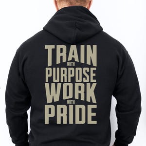 May include: A black hooded sweatshirt featuring large, light-colored block lettering on the back that reads "TRAIN WITH PURPOSE WORK WITH PRIDE." The text is arranged in four lines, with the words "WITH" appearing in smaller font between the main lines.