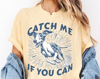 May include: A pale yellow short-sleeve t-shirt featuring a dark blue graphic of a running greyhound dog. The text "CATCH ME IF YOU CAN" is printed in dark blue capital letters above and below the dog illustration, which includes radiating lines.