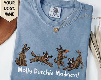 May include: A blue short-sleeve t-shirt featuring a graphic of four brown dogs in various poses, including running and playing with a frisbee. The text "Molly Dutchie Madness!" is printed in white below the dogs. A gold-tone necklace rests on the collar.