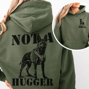 Puede incluir: Sudadera con capucha verde oliva con un gráfico negro de un perro con gafas y arnés. El texto "NOTA HUGGER" está impreso debajo del perro. Un gráfico más pequeño del perro y el nombre "BROLIN" están en la parte superior derecha.