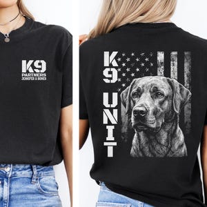 May include: A black t-shirt featuring a white graphic on the front chest that reads K9 PARTNERS JENNIFER & BONES. The back displays a large white graphic of a dog, the text K9 UNIT, and a distressed American flag design in a monochromatic style.