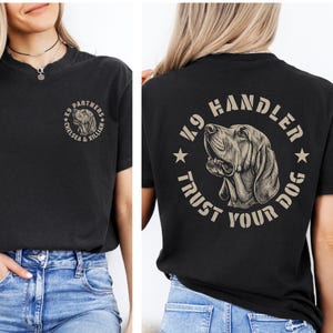 Puede incluir: Una camiseta negra con un pequeño gráfico circular en el pecho izquierdo que muestra una ilustración de un perro y el texto K9 PARTNERS CHELSEA & KILLIAN. La parte trasera muestra un gráfico circular grande con un perro y el texto K9 HANDLER TRUST YOUR DOG.