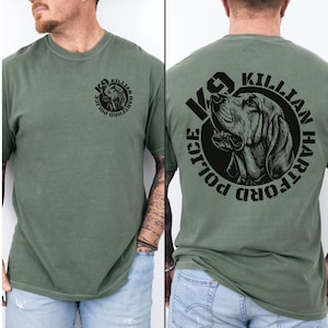Puede incluir: Una camiseta de manga corta color verde oliva con un gráfico circular negro en el pecho izquierdo y una versión más grande en la espalda. El gráfico muestra una cabeza de perro con el texto K9 KILLIAN HARTFORD POLICE en un borde circular.