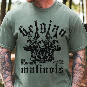 Op de afbeelding: Een saliegroen T-shirt met korte mouwen en een zwarte afbeelding van drie koppen van een Belgische Mechelse herder. De tekst luidt: Belgian, K9 GUNNER, Not a Pet. A Partner. en malinois. Het ontwerp staat centraal op de borst van het shirt.