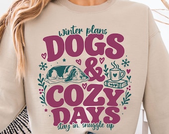 May include: A beige crewneck sweatshirt featuring the dark pink text "DOGS & COZY DAYS" and dark green text "winter plans" and "stay in, snuggle up". The graphic includes a sleeping dog, a steaming mug on a book, hearts, and snowflakes.