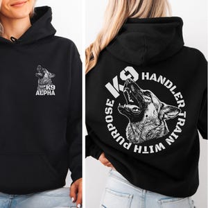 May include: A black hooded sweatshirt featuring a white graphic of a German Shepherd on the front left chest with the text K9 ALPHA. The back displays a large circular graphic of a German Shepherd with the text K9 HANDLER TRAIN WITH PURPOSE.