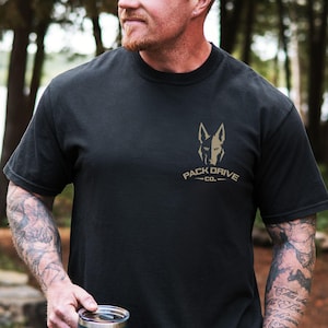 Puede incluir: Una camiseta negra de manga corta y cuello redondo con un gráfico de color tostado en el pecho izquierdo. El diseño muestra la silueta estilizada de una cabeza de perro sobre el texto PACK DRIVE CO. en una fuente negrita con líneas laterales.